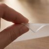 ANRO_mattierte_Folie_2mm_08-jpg Hand hält matte, 2mm dicke transparente Tischschutzfolie über einem Holztisch.